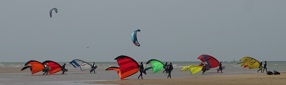 Image d'une groupe de gens qui traverse une plage de sable fin vers la mer en portant les volants de kitesurf